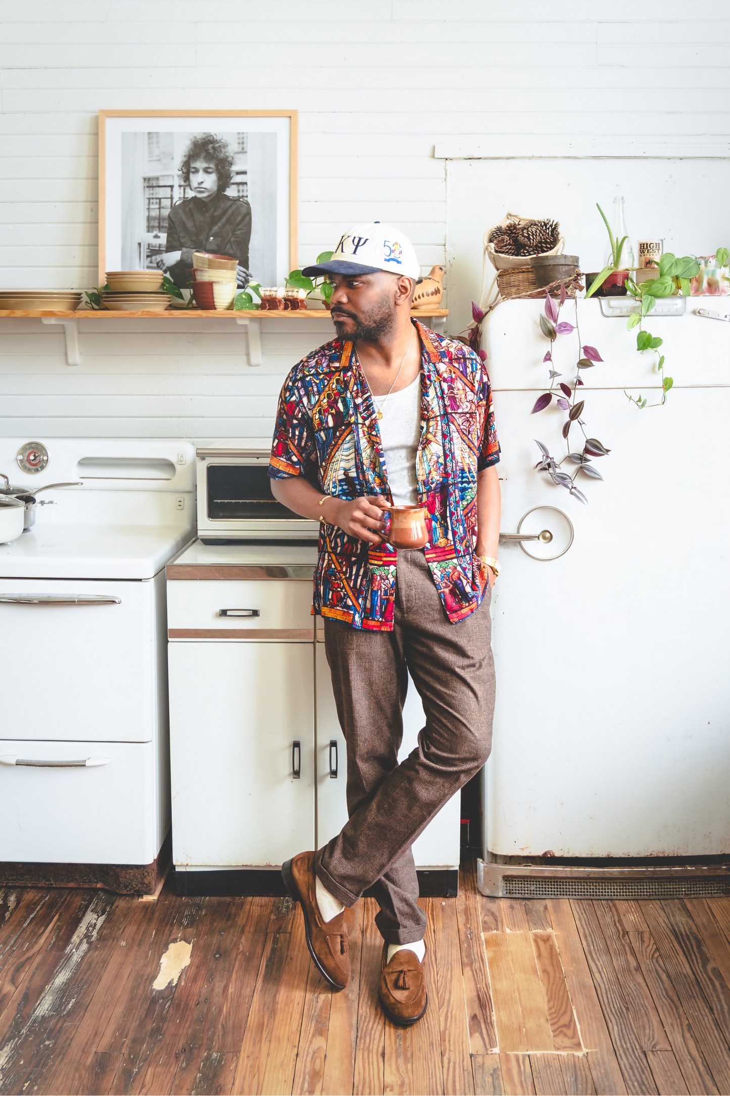 A male model wearing a camp collar shirt with a pattern inspired by the Tuskegee University Chapel's "singing windows" stained glass installation, standing in a room with light-colored walls.