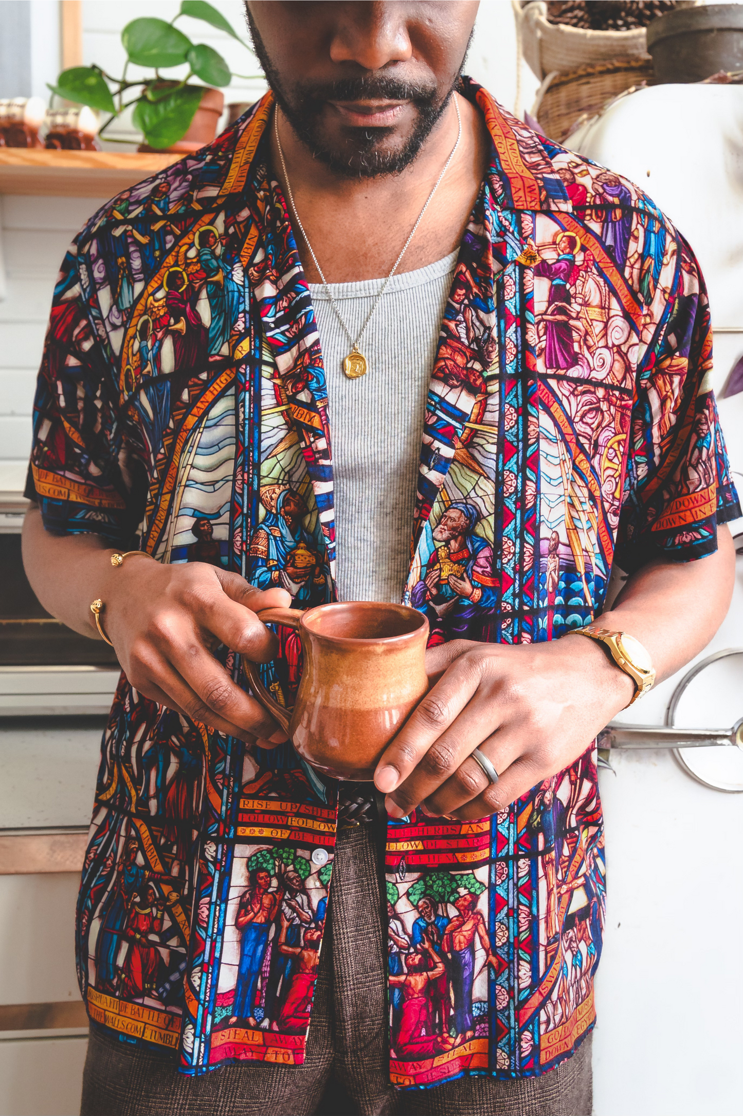 A male model wearing a camp collar shirt with a pattern inspired by the Tuskegee University Chapel's "singing windows" stained glass installation, standing in a room with light-colored walls.