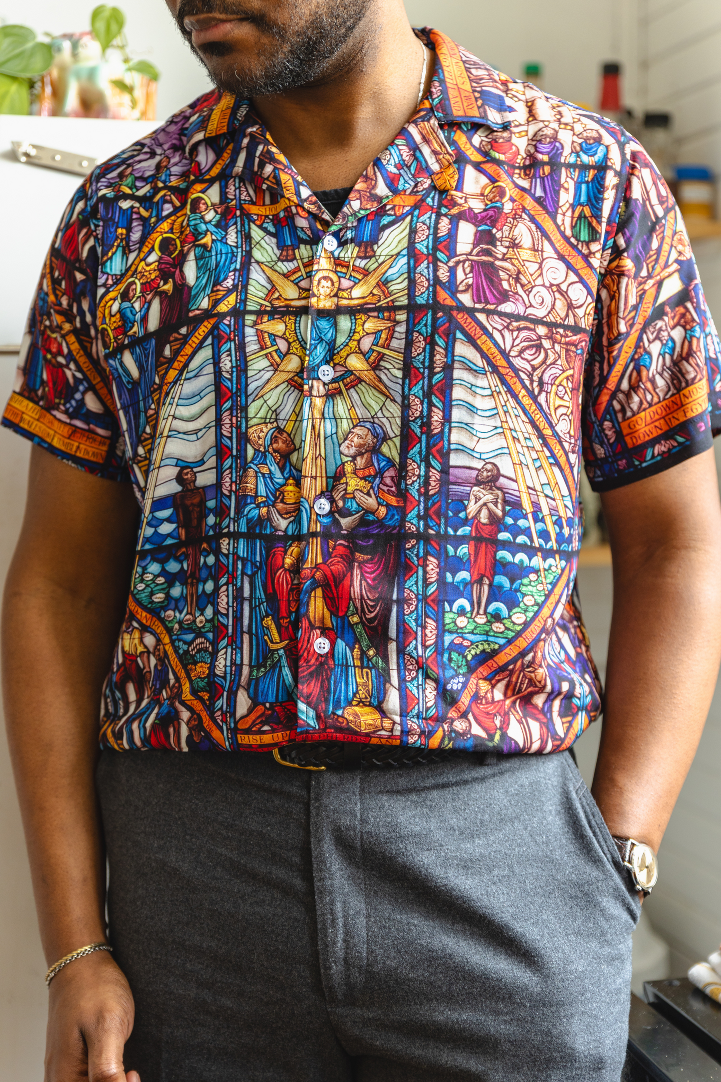 A male model wearing a camp collar shirt with a pattern inspired by the Tuskegee University Chapel's "singing windows" stained glass installation, standing in a room with light-colored walls.
