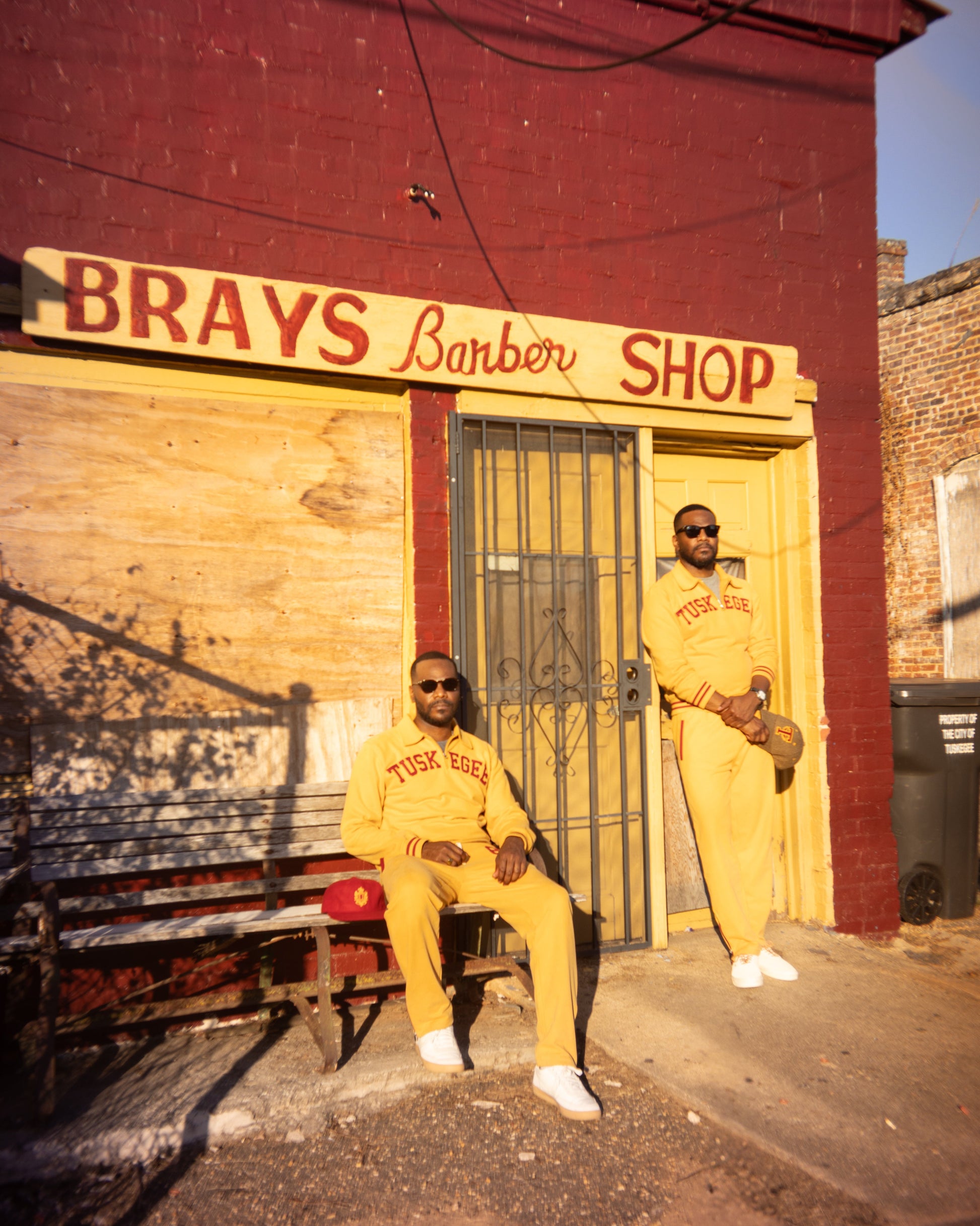 Two men wearing matching yellow tracksuits with 'Tuskegee' printed on the chest pose in front of 'Brays Barber Shop' in Tuskegee Alabama